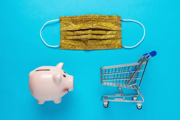 Piggy bank with a medical face mask bancnote and shopping cart on blue background. Minimal Coronavirus outbreak. Saving money concept.
