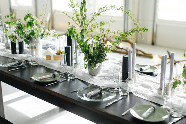 Stylish meals and a long table, loft. Black table, chairs, dishes, candles. Banks with greens, flowers. Black candles