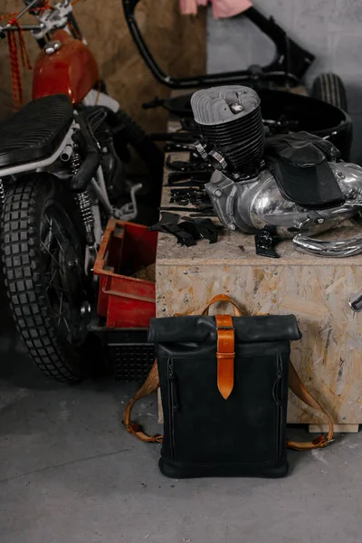 Exclusive black leather backpack. Backpack near the wall. The tools in the garage. Many different tools. Motorcycle in the garage.