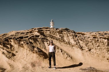 adam sahilde. Kumlu kayalıklardan yakınındaki bir adam. Adam içinde belgili tanımlık geçmiş deniz feneri. Deniz feneri