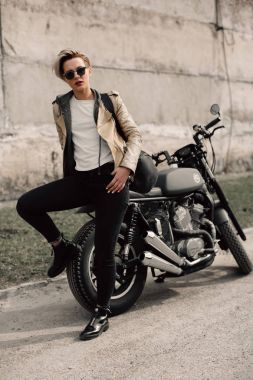 Onun motosiklet üzerinde oturan bir bayan. Gri bir duvara yakın motosiklet. kısa saçlı kız. kız siyah gözlük ve bir deri ceket. Bisiklet