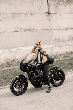 Onun motosiklet üzerinde oturan bir bayan. Gri bir duvara yakın motosiklet. kısa saçlı kız. kız siyah gözlük ve bir deri ceket. Bisiklet