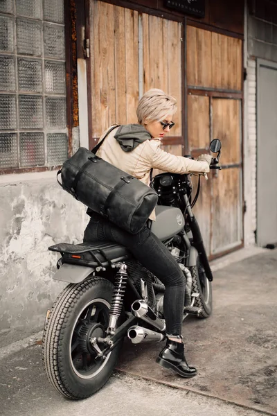 Femme assise sur sa moto. Moto près du garage. Une femme vêtue de cuir. Moto noire. Une femme avec une coupe courte. Vélo — Photo