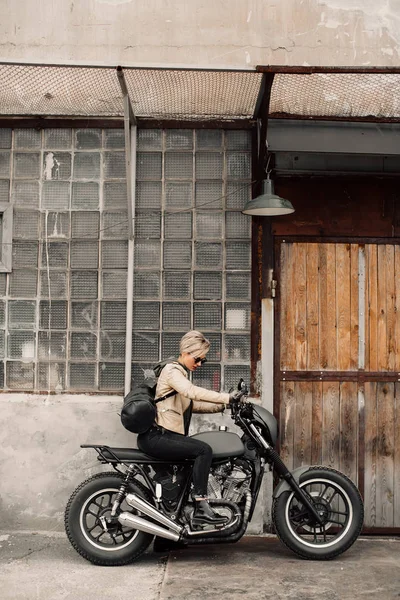 Mujer sentada en su motocicleta. Motocicleta cerca del garaje. Una mujer con una chaqueta de cuero. Moto negro. Una mujer con un corte de pelo corto. Moto —  Fotos de Stock