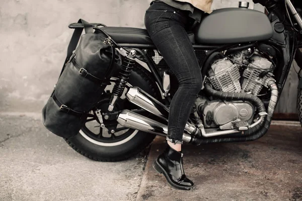 Γυναίκα που κάθεται στη μοτοσικλέτα του. Μοτοσικλέτα κοντά στο γκαράζ. Δερμάτινη τσάντα και τζιν. Μοτοσικλέτα μαύρη. Στα πόδια της γυναίκας σε μια μοτοσικλέτα. Ποδήλατο — Φωτογραφία Αρχείου