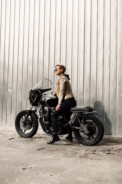 Woman sitting on her motorcycle. Motorcycle near a grey wall. girl with short hair. girl in black glasses and a leather jacket. Bike