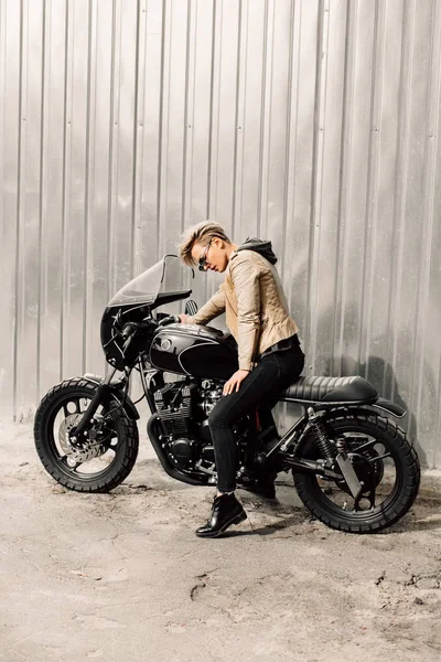 Mujer sentada en su motocicleta. Moto cerca de una pared gris. chica con el pelo corto. chica en gafas negras y una chaqueta de cuero. Moto —  Fotos de Stock