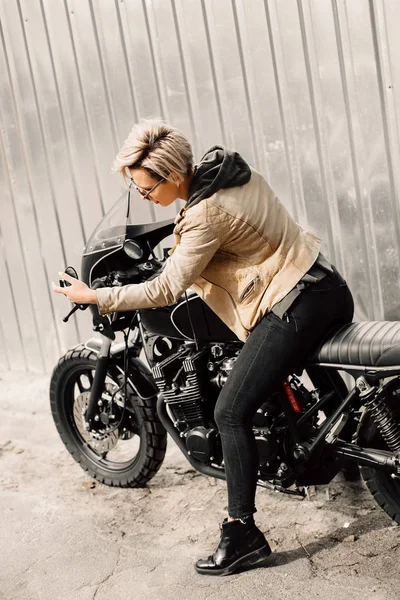 Femme assise sur sa moto. Moto près d'un mur gris. fille se regarde dans le miroir de la moto. fille aux cheveux courts. fille en lunettes noires et une veste en cuir. Vélo — Photo