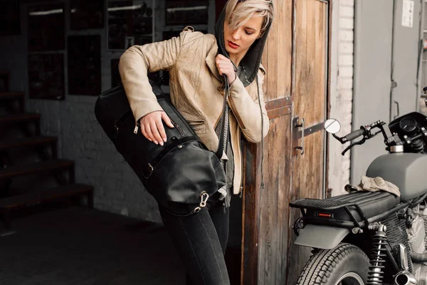 Una joven con una bolsa de cuero cerca del garaje. Una mujer con una chaqueta de cuero. Motocicleta cerca del garaje. Bolso de cuero Imagen De Stock