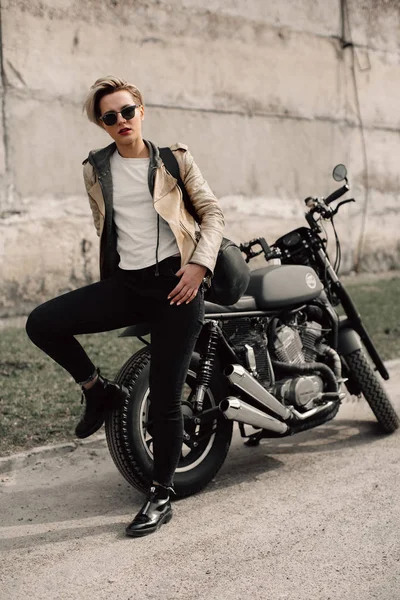 Mujer sentada en su motocicleta. Moto cerca de una pared gris. chica con el pelo corto. chica en gafas negras y una chaqueta de cuero. Moto Imagen De Stock