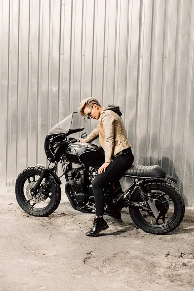 Mujer sentada en su motocicleta. Moto cerca de una pared gris. chica con el pelo corto. chica en gafas negras y una chaqueta de cuero. Moto Imagen De Stock