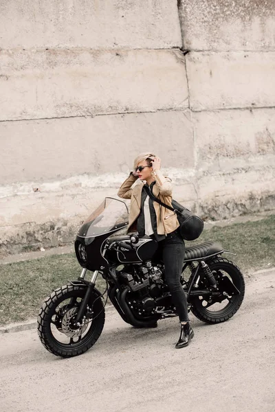 Mujer sentada en su motocicleta. Moto cerca de una pared gris. chica con el pelo corto. chica en gafas negras y una chaqueta de cuero. Moto Imágenes De Stock Sin Royalties Gratis