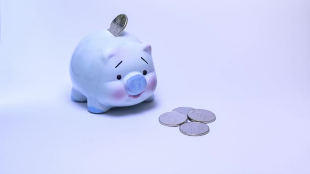 Banco piggy azul claro com muitas moedas de dinheiro em fundo branco e ideias de poupança de dinheiro. Prazo de validade 4K — Vídeo de Stock
