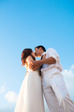 Romantic couple dressed in white, kissing under the blue sky on their wedding day. clipart