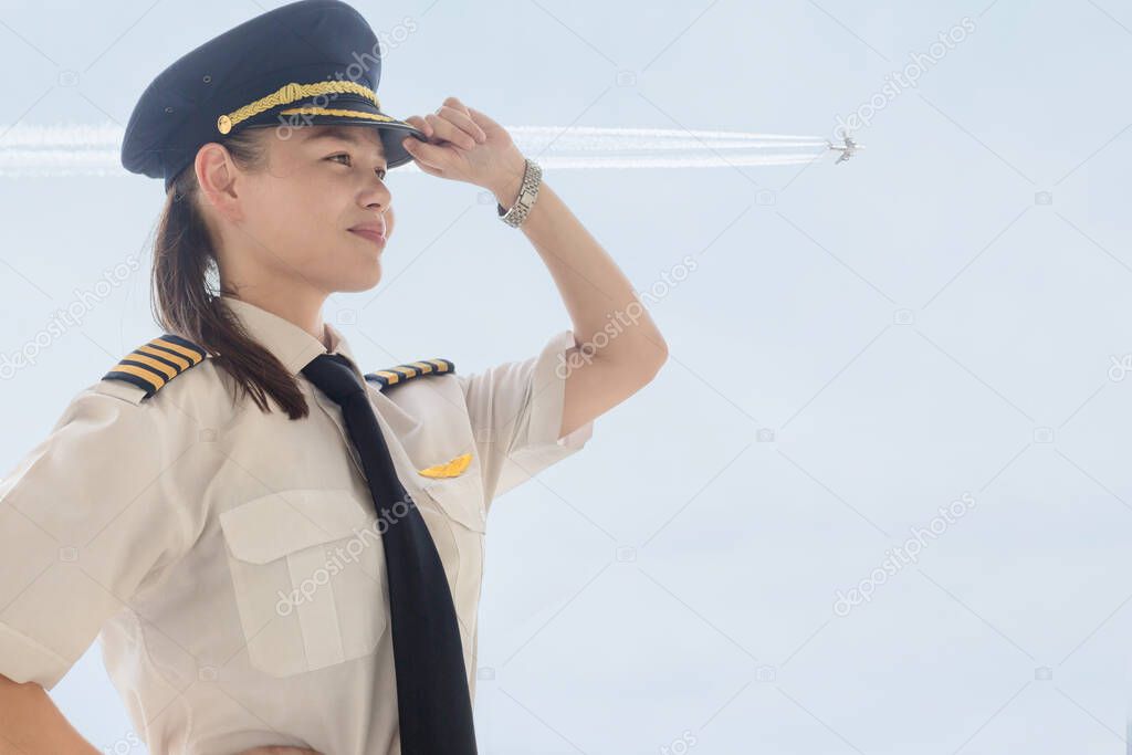 Sexy professional pilot woman with hat on saluting at the airport with a airplane contrail in the sky.