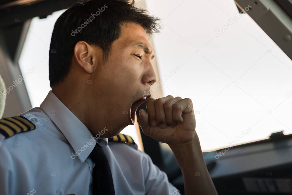 A male captain pilot sitting in the cockpit yawning while flying an airplane