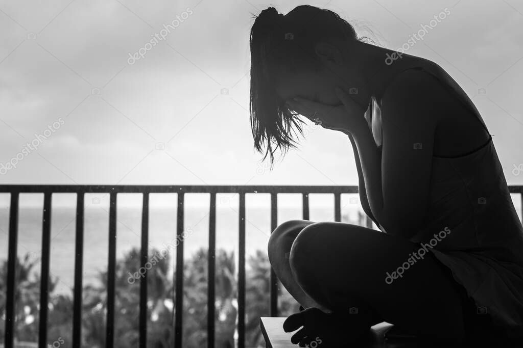 Silloutte of a woman sitting alone on her hotel balcony weeping and covering her face, black and white.