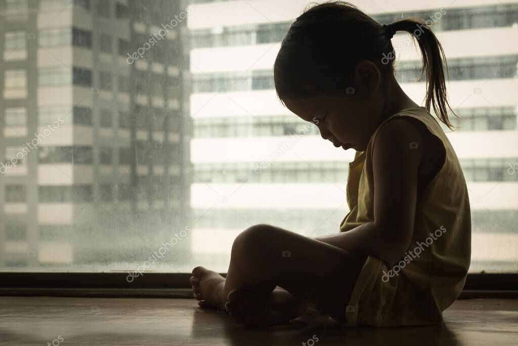 A litte girl sitting next to a window with her head down in sadness. Feeling depressed and hurt.