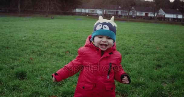 Glada lilla pojken i röd jacka kör med leende i park. Underordnad som körs på grönt gräs. Slow motion 4k. — Stockvideo