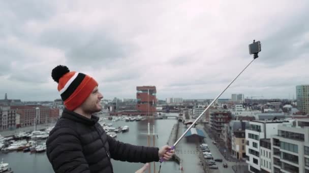 Jovem faz selfie no telhado com vista para a paisagem urbana. Sentimento de felicidade. 4K. Em chapéu colorido . — Vídeo de Stock