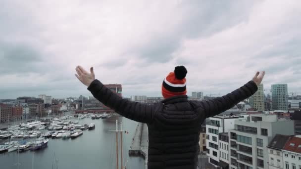 Man in special coloring hat, standing on barrier on roof, spreading wide his hands, looking into distance. 4k. Feeling of happiness. — Stock Video