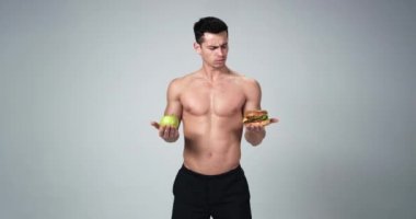 Atletik kaslı adam elinde tutan bir hamburger ve bir elma, sağlıklı bir yaşam tarzı olur. 4k