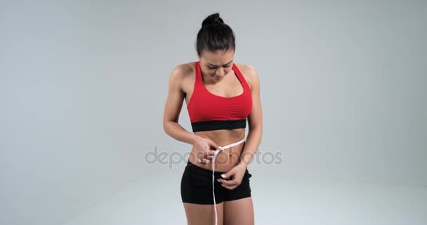 年轻女运动健身上衣和短裤持有测量与磁带的腹部，她微笑和孤立的白色背景上。4 k.慢动作 — 图库视频影像