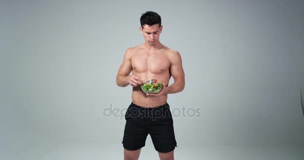プラスチック製のボウルを持って、白い背景と彼の笑顔は、健康的な生活のために、新鮮なサラダを食べる男性フィットネス モデル。4 k. スローモーション — ストック動画