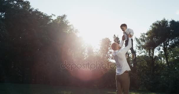 O pai novo que cria a criança no ar sobre a cabeça na natureza, papai e filho jogam no parque ao ar livre no dia de verão. Pôr do sol sobre o fundo. MoTion lento — Vídeo de Stock