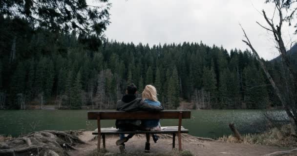 Pareja joven sentada en el banco de madera. Miré el lago. Mirando enamorados el uno del otro — Vídeo de stock