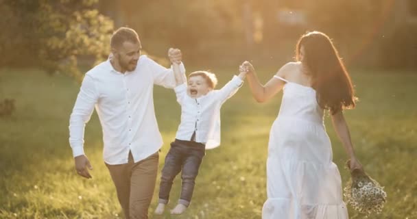 Joven familia feliz con el bebé. Van al pintoresco campo verde al atardecer. El niño es criado por los padres. Puesta de sol sobre el fondo . — Vídeo de stock