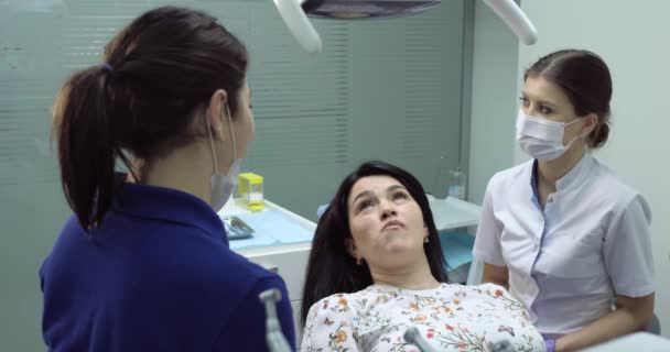 Стоматолог проверяет зубы молодой женщины с зеркалом, зубная медсестра сидит рядом и помогает, пациент счастлив открыть рот — стоковое видео