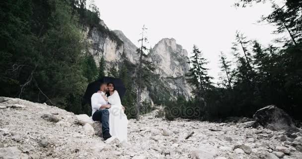 Schönes junges Paar bleibt auf dem Felsen unter dem Regenschirm umarmt einander, Hintergrund erstaunliche Landschaft. — Stockvideo