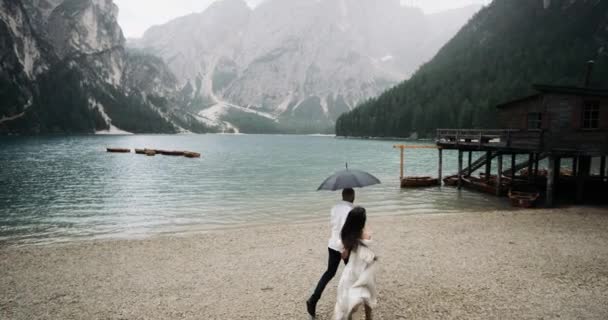Fantastická krajina s velkým jezerem a horami v den déšť s pár běžící pod deštníkem do dřevěného domu. Zpomalený pohyb. 4 k. Red Epic. Lago de Braies — Stock video