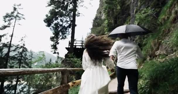 Casal jovem na viagem romântica que corre da ponte de madeira no meio da paisagem surpreendente com grandes montanhas. movimento desleixado — Vídeo de Stock