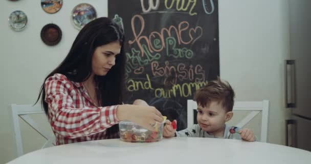 Madre con su pequeño hijo sentado en la silla de la cocina jugando con gelatina y besándose — Vídeos de Stock