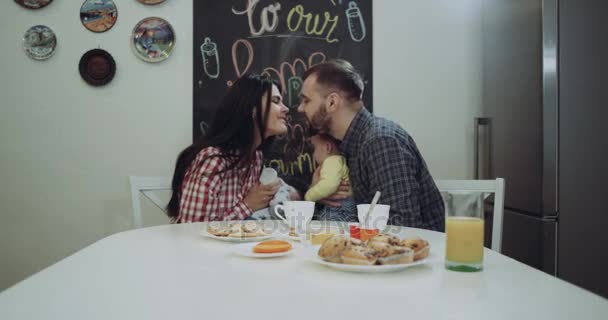 Familia joven y dos niños pequeños pasando la mañana juntos en la mesa de la cocina y felices se besan . — Vídeo de stock