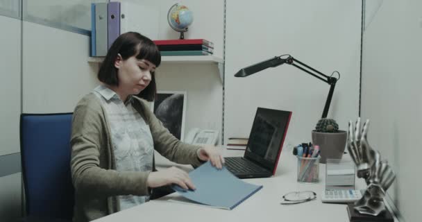 Trabajadora de oficina sentada en su escritorio mirando el papeleo y verificando en su cuaderno algo de trabajo . — Vídeos de Stock