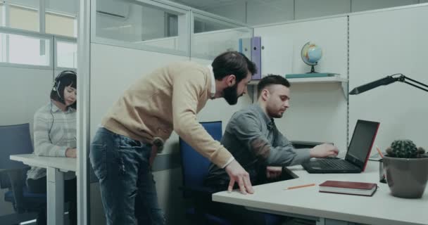 办公室经理站在桌子旁边, 用膝上型电脑向他的办公室工作人员解释一些事情, 后台一位客户支持工作人员坐在她的办公桌上. — 图库视频影像