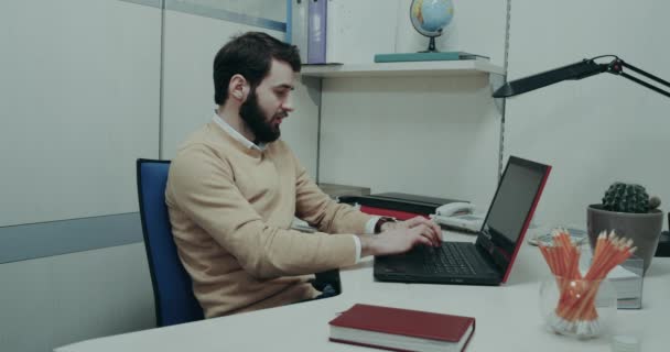 Ffice 的工人, 坐在他的办公桌上敲击他的笔记本电脑键盘, 有一个集中和微笑的脸. — 图库视频影像
