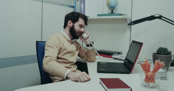 Pracownik biurowy, siedząc na jego stole działa za pośrednictwem laptopa zmęczony i nerwowo mówiąc na telefon — Wideo stockowe