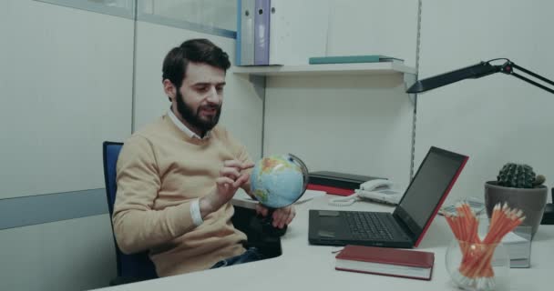 Escritório jogando carismático com um clipart geográfico em sua mesa de trabalho, em seguida, começar a trabalhar em seu laptop — Vídeo de Stock