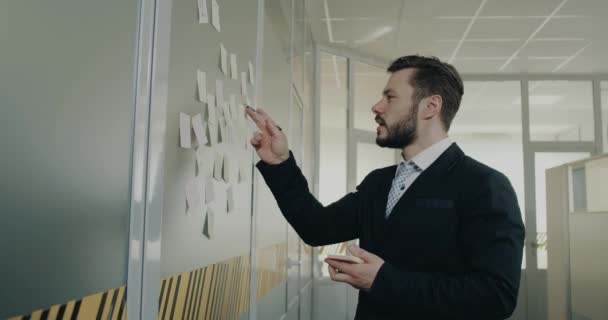 若いビジネスの男性が小さな紙、オフィスの壁に棒にいくつかの計画を書く — ストック動画