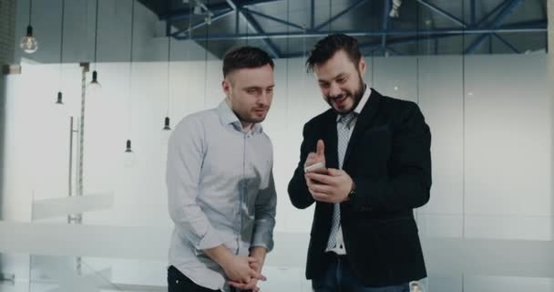 Deux jeunes collègues ont une conversation dans le couloir du bureau regardant le téléphone curieusement — Video