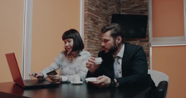 Two colleagues in the cafe sitting at the coffee table and using the card and laptop buy something — Stock Video