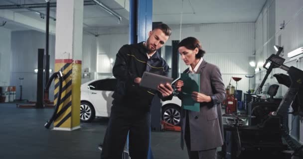 W centrum usług samochodowych dobrze wyglądająca kobieta w garniturze analizując problem samochodu z jej mechanikiem następnie spojrzeć na samochód — Wideo stockowe