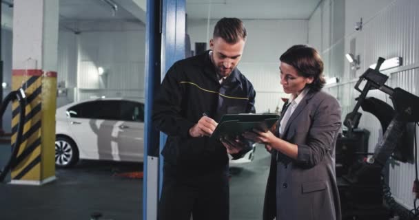 In einem großen Service-Auto-Center charismatischer Mechaniker Mann in Uniform mit einer Geschäftsfrau unterschreiben einige Papiere dann kommen sie neben dem Auto, um das Problem zu überprüfen — Stockvideo