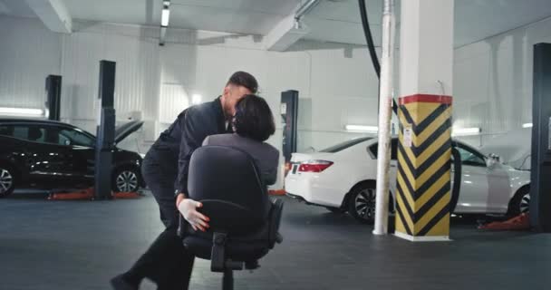 Δύο αστείοι εργαζόμενοι μηχανικός και διευθυντής γυναίκα σε ένα κοστούμι έχουν ένα διάλειμμα χρόνο μπροστά από την κάμερα σε ένα κέντρο παροχής υπηρεσιών auto — Αρχείο Βίντεο