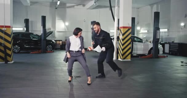 Femme gestionnaire élégant dans un costume et un mécanicien de gars dans un service auto danse excité devant la caméra dans une voiture de service moderne — Video
