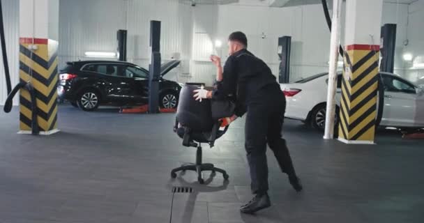 Good looking guy and his manager woman have a fun time together in a service auto center they play on a chair dancing — Stock Video